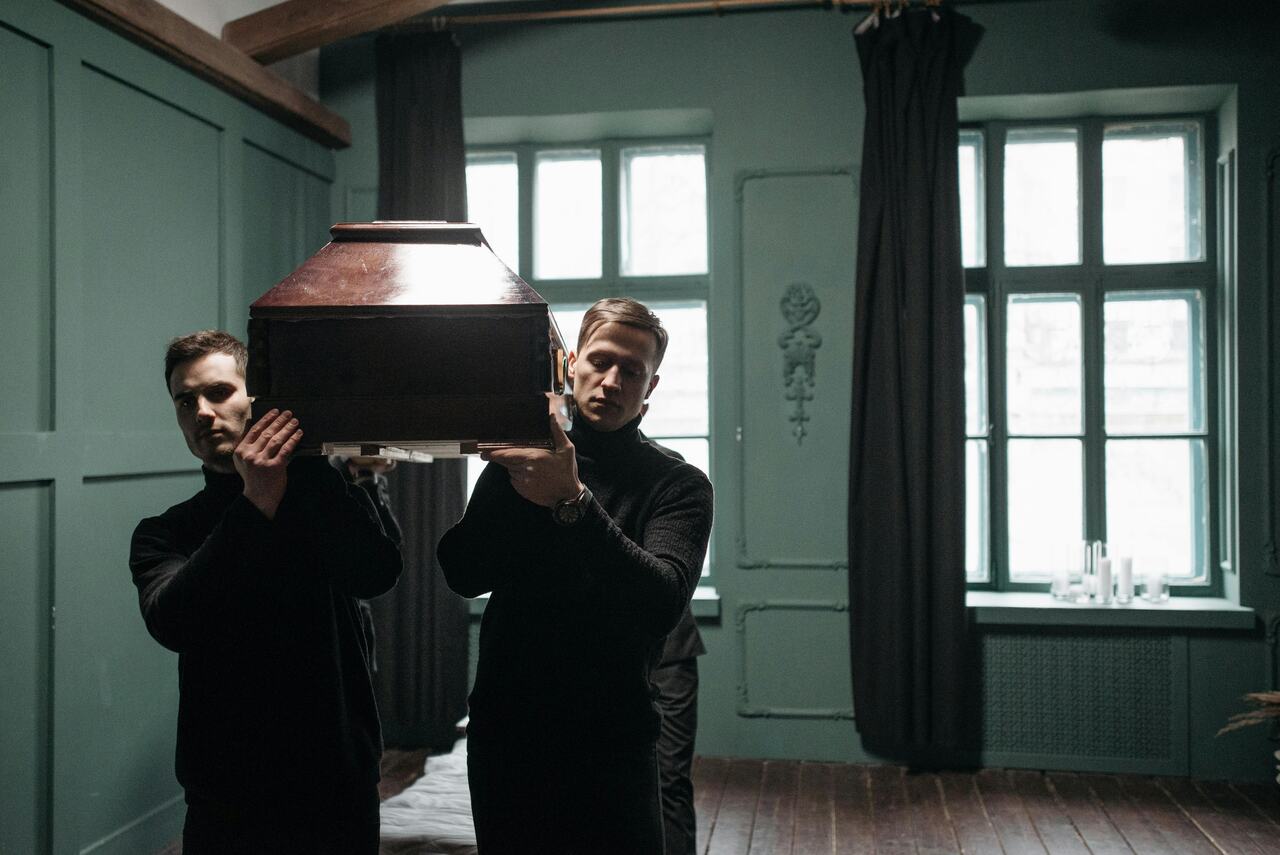 Dois homens carregando um caixão de madeira dentro de uma sala de velório com janelas grandes e cortinas escuras.