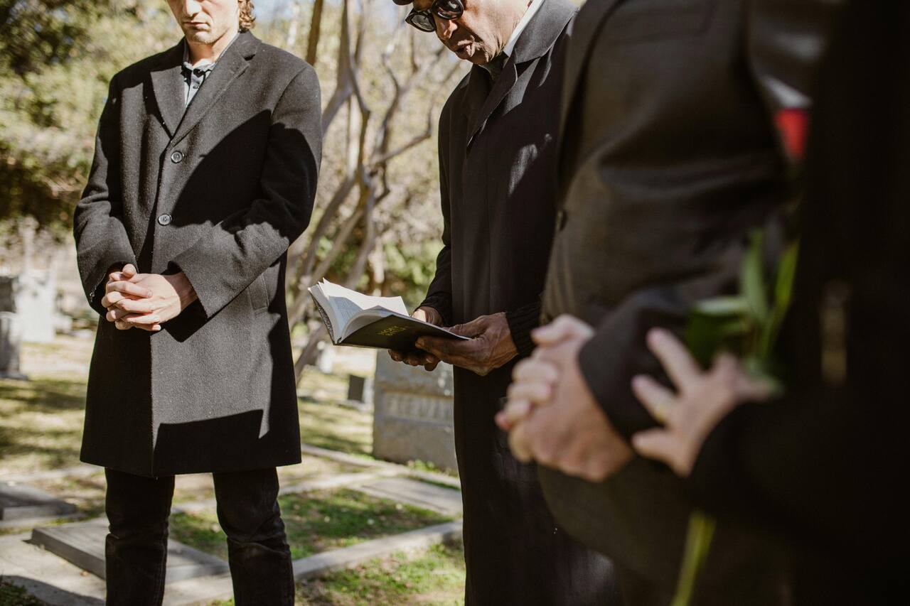 Grupo de pessoas em um sepultamento, com um homem lendo um livro de orações enquanto os demais mantêm as mãos cruzadas em respeito.