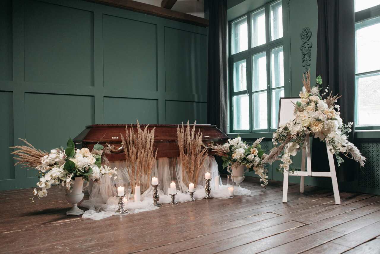 Caixão de madeira em uma sala de velório decorada com arranjos florais brancos, velas e elementos rústicos.