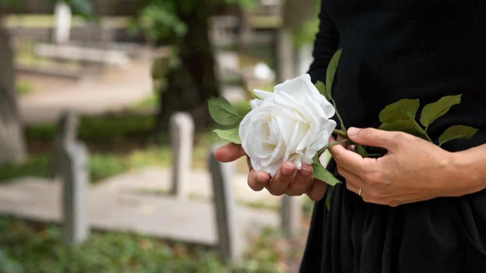 Mãos segurando uma rosa branca durante visita a um cemitério, imagem associada ao auxílio funeral em momentos de perda.